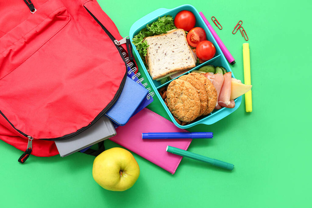 Рюкзак, канцелярские принадлежности и обеденная коробка с вкусной едой на зеленом фоне - Фото, изображение