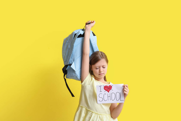 Little girl holding paper with text I DO NOT LOVE SCHOOL and backpack on yellow background - Photo, Image
