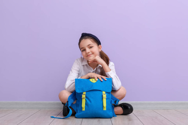 Pequena estudante com mochila sentada perto da parede lilás - Foto, Imagem