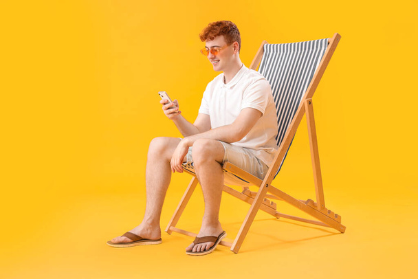 Jovem usando telefone celular em cadeira de praia no fundo amarelo - Foto, Imagem