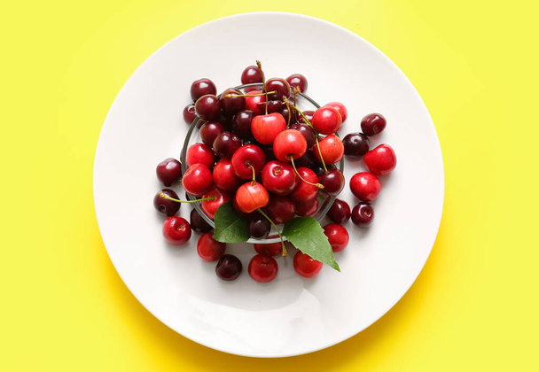 Plate and glass bowl with sweet cherries on yellow background - 写真・画像
