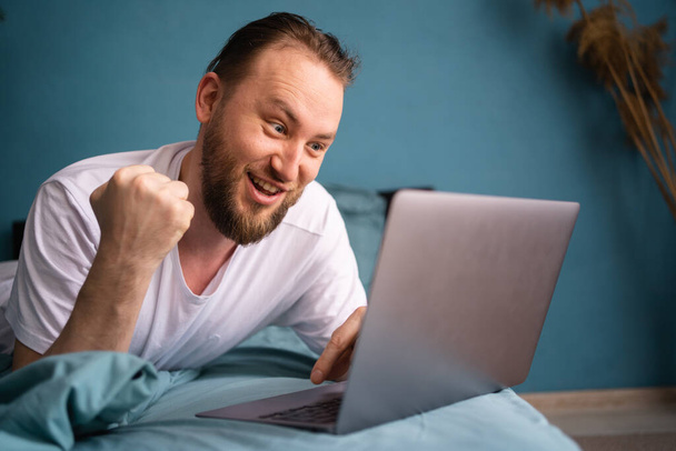 Portrait of happy man spending his weekend laying in bed, watching soccer play live broadcast on his laptop and celebrating money win from bets on his favourite team. Close-up - Фото, зображення
