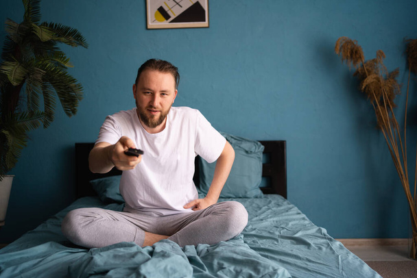 Homem sem sono no pijama assistindo tv enquanto sentado na cama com controle remoto. Espaço de cópia - Foto, Imagem