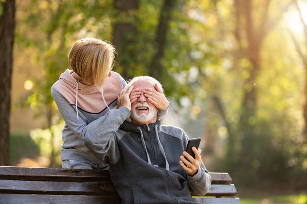 starsza para korzystająca z telefonu komórkowego siedząca na ławce w parku - Zdjęcie, obraz