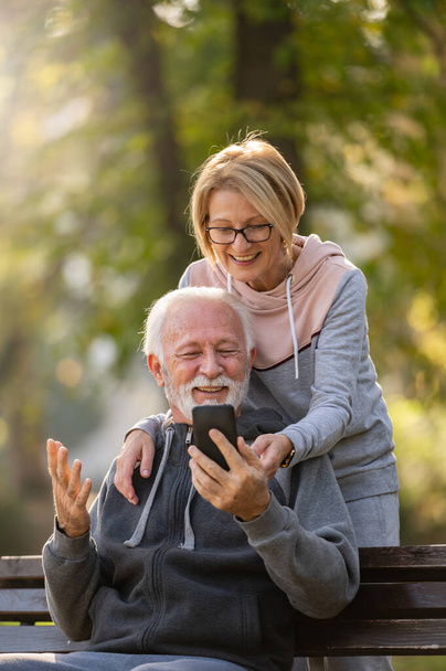 Älteres Ehepaar sitzt mit Handy auf Bank im Park - Foto, Bild