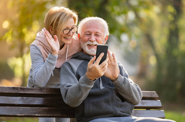 ηλικιωμένο ζευγάρι που χρησιμοποιεί κινητό τηλέφωνο κάθεται στον πάγκο στο πάρκο - Φωτογραφία, εικόνα