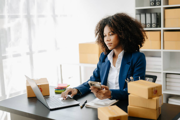 Startup small business SME, Entrepreneur owner African woman using laptop taking receive and checking online purchase shopping order to prepare pack product boxes in office - 写真・画像