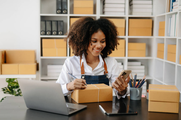 Startup small business SME, Entrepreneur owner African woman using laptop taking receive and checking online purchase shopping order to prepare pack product boxes in office - Φωτογραφία, εικόνα