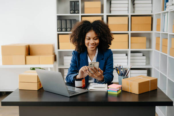 Startup small business SME, Entrepreneur owner African woman using laptop taking receive and checking online purchase shopping order to prepare pack product boxes in office - 写真・画像