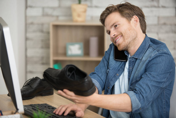 man on computer holding shoe and talking on smartphone - Φωτογραφία, εικόνα