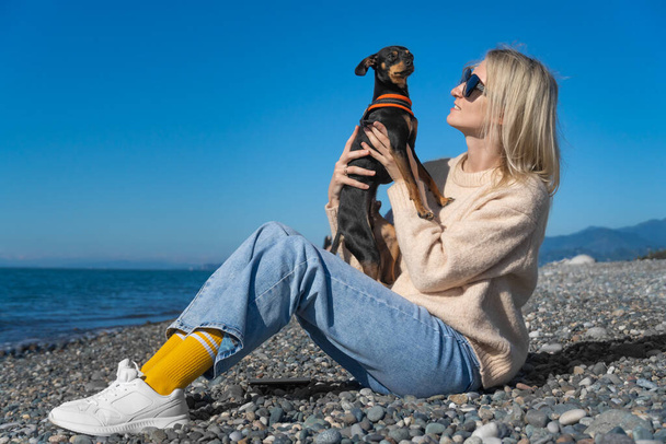 Młoda piękna blondynka w okularach przeciwsłonecznych siedzi na wybrzeżu w słoneczny dzień i trzyma w ramionach rosyjskiego psa Toy Terrier. Przyjaźń między psem a mężczyzną. - Zdjęcie, obraz
