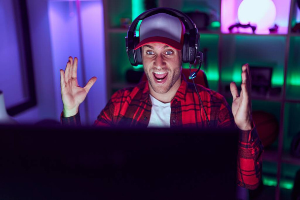 Young caucasian man playing video games celebrating victory with happy smile and winner expression with raised hands  - Фото, изображение