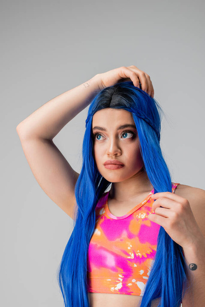 self expression, young woman adjusting blue hair and looking away on grey background, isolated, fashion choices, stylish look, colorful clothes, casual attire, generation z fashion, long hair - Foto, immagini