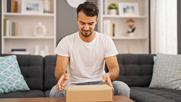 Mladý hispánec muž vybalování lepenkové krabice sedí na pohovce doma - Fotografie, Obrázek