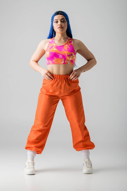 fashion, tattooed young woman with blue hair posing with hands on hips and standing in colorful clothes on grey background, full length, individualism, modern style, urban fashion, vibrant color - Foto, afbeelding