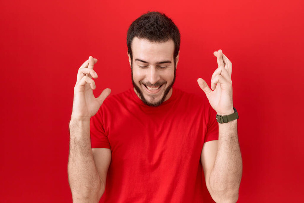 Young hispanic man wearing casual red t shirt gesturing finger crossed smiling with hope and eyes closed. luck and superstitious concept.  - Photo, Image