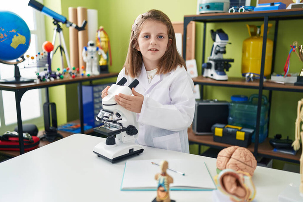 Suloinen blondi tyttö opiskelija hymyilee luottavainen käyttäen mikroskooppi laboratoriossa luokkahuoneessa - Valokuva, kuva