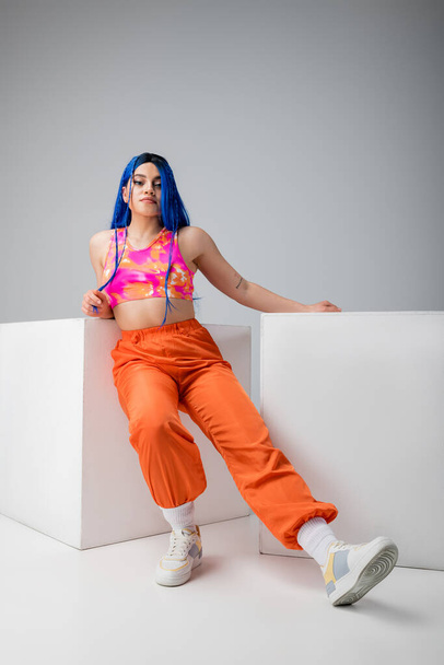 modern subculture, tattooed woman with blue hair posing in vibrant clothes near white cubes on grey background, full length, individualism, modern style, urban fashion, young model  - Foto, Imagen