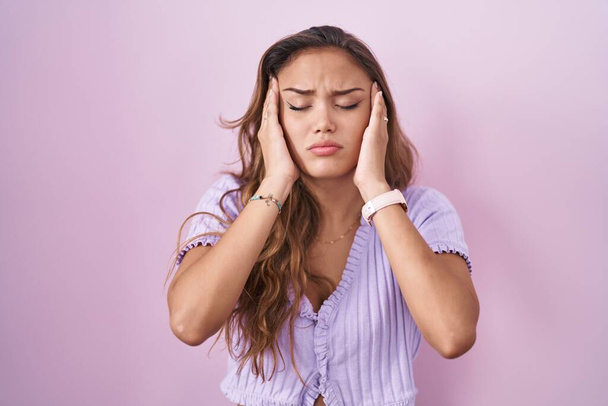 Fiatal spanyol nő áll a rózsaszín háttér felett, kéz a fejen, fejfájás, mert a stressz. migrénben szenved.  - Fotó, kép