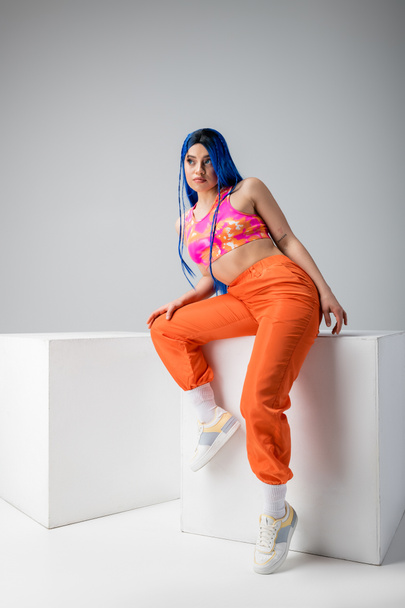 urban fashion, tattooed woman with blue hair posing in colorful clothes near white cubes on grey background, full length, individualism, modern style, vibrant color, young model  - Photo, Image