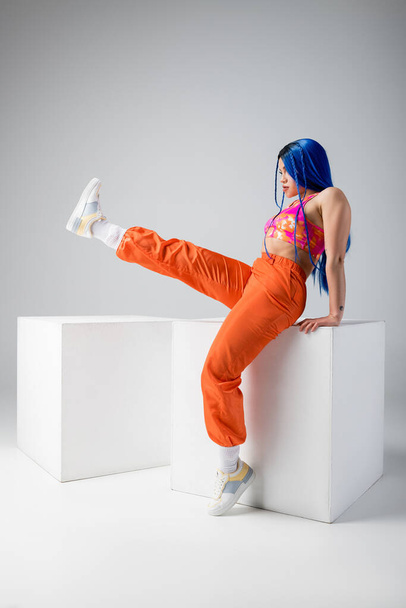 fashion trends, tattooed young woman with blue hair posing with raised leg near white cubes on grey background, full length, individualism, modern style, urban fashion, vibrant color, model  - Foto, Imagen
