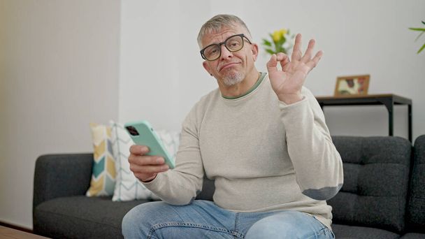 Keski-ikä harmaa tukka mies käyttää älypuhelin tekee ok ele kotona - Valokuva, kuva