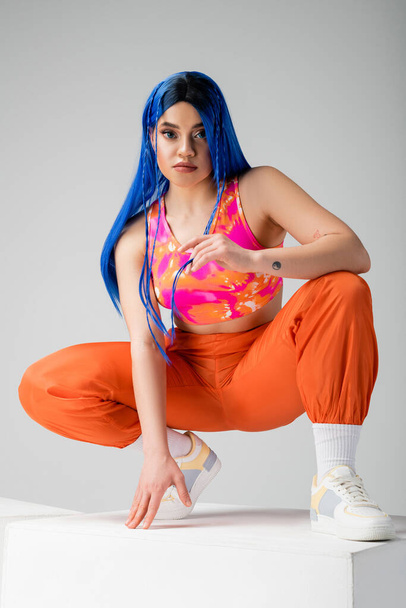 youthful fashion, tattooed young woman with blue hair sitting on top of white cube on grey background, full length, individualism, modern style, urban fashion, vibrant color, model  - Φωτογραφία, εικόνα