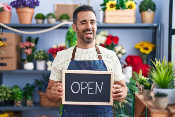 Hispanic man with beard working at florist holding open sign winking looking at the camera with sexy expression, cheerful and happy face.  - Photo, Image
