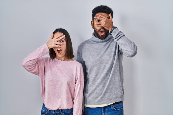 Young hispanic couple standing together peeking in shock covering face and eyes with hand, looking through fingers with embarrassed expression.  - Photo, image