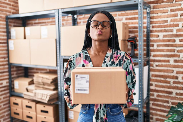 African woman with braids working at small business ecommerce holding package looking at the camera blowing a kiss being lovely and sexy. love expression.  - 写真・画像