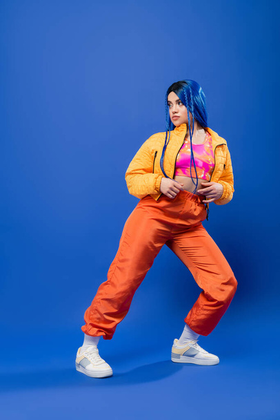 full length, dyed hair, fashion forward, tattooed female model with blue hair posing in puffer jacket and orange pants on blue background, vibrant color, urban fashion, individualism, young woman  - Photo, Image