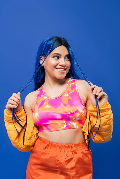 beauty trends, dyed hair, cheerful female model with blue hair posing in puffer jacket on blue background, vibrant color, urban fashion, individualism, young woman smiling and looking away - Foto, imagen