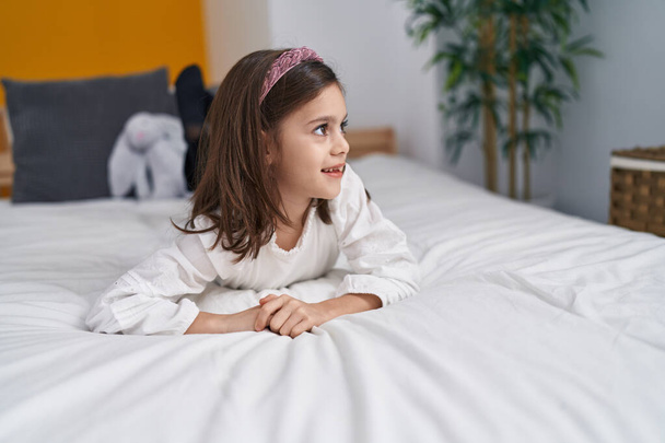 Liebenswertes hispanisches Mädchen lächelt selbstbewusst auf dem Bett im Schlafzimmer liegend - Foto, Bild