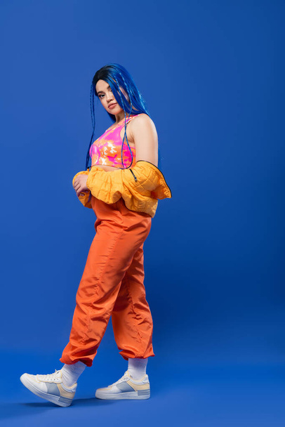 longitud completa, mujer tatuada con el pelo teñido, la moda hacia adelante, modelo femenino con el pelo azul posando en chaqueta hinchable y pantalones de color naranja sobre fondo azul, color vibrante, moda urbana, individualismo  - Foto, imagen