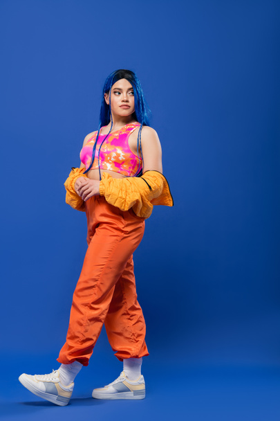 comprimento total, mulher com cabelo tingido, declaração de moda, modelo feminino tatuado com cabelo azul posando na jaqueta puffer e calças laranja no fundo azul, cor vibrante, moda urbana, individualismo  - Foto, Imagem
