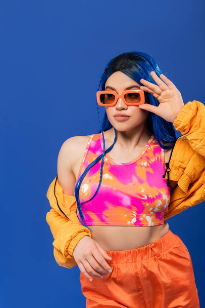 fashion statement, young female model with blue hair adjusting trendy sunglasses isolated on blue background, generation z, rebel style, colorful clothes, individualism, modern woman  - 写真・画像