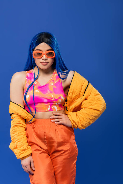 trendy accessory, young female model with blue hair and trendy sunglasses posing with hand on hip isolated on blue background, rebel style, colorful clothes, individualism, modern woman  - Foto, Bild