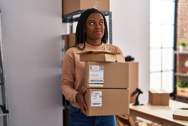 Young african american with braids working at small business ecommerce holding packages smiling looking to the side and staring away thinking.  - Foto, Imagem