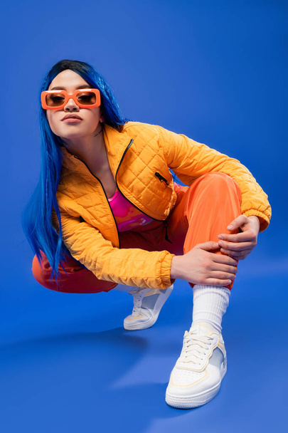 full length of pretty young female model with blue hair and trendy sunglasses sitting on haunches on blue background, generation z, rebel style, individualism, modern fashion, trendy accessory  - Φωτογραφία, εικόνα