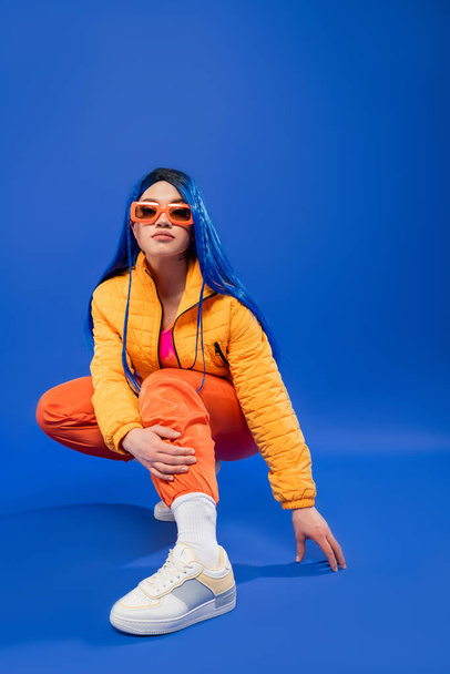 individualism, full length of pretty female model with blue hair and trendy sunglasses sitting on haunches on blue background, rebel style, modern fashion, trendy accessory, generation z,  - Foto, imagen
