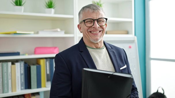Middle age grey-haired man teacher smiling confident holding folder at university classroom - 写真・画像