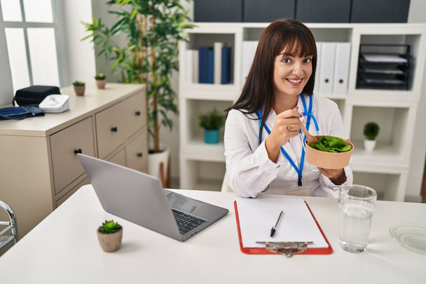 Junge schöne hispanische Ärztin mit Laptop Salat essen in Klinik - Foto, Bild