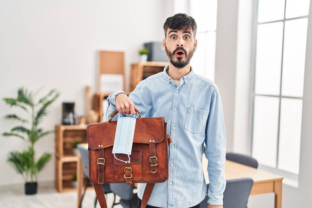 Junger hispanischer Mann mit Bart, der im Büro mit Koffer und Schutzmaske arbeitet, erschrocken und erstaunt mit offenem Mund für Überraschung, ungläubiges Gesicht  - Foto, Bild