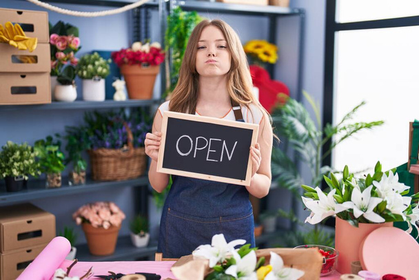 Caucasian woman working at florist holding open sign puffing cheeks with funny face. mouth inflated with air, catching air.  - Photo, image