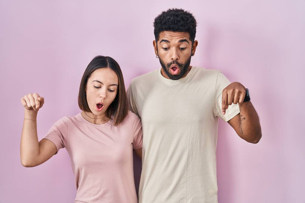 Mladý hispánský pár spolu přes růžové pozadí ukazuje dolů s prsty zobrazující reklamu, překvapený obličej a otevřená ústa  - Fotografie, Obrázek
