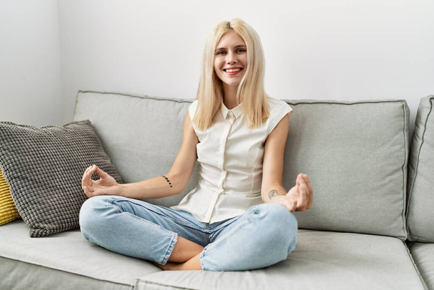Jeune femme blonde faisant de l'exercice de yoga assis sur le canapé à la maison - Photo, image