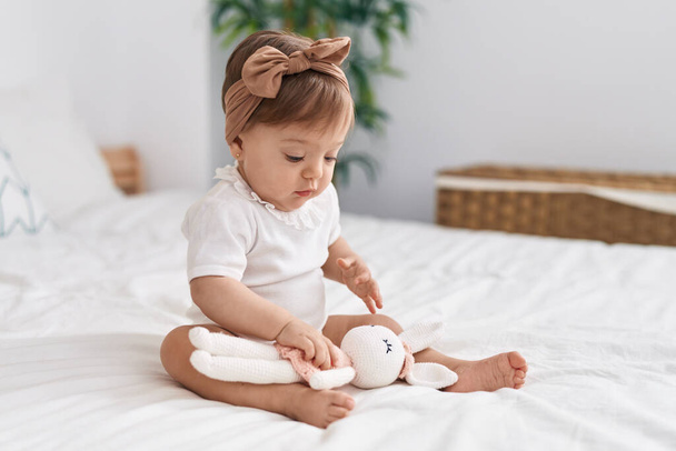 Adorable caucasian baby sitting on bed playing with doll at bedroom - Foto, imagen