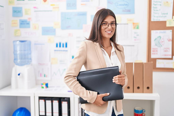 Young beautiful hispanic woman business worker smiling confident holding binder at office - Photo, Image