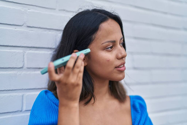 Junge hispanische Frau hört Audiobotschaft mit dem Smartphone über weißen isolierten Backsteinhintergrund - Foto, Bild