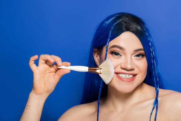 beauty trends, individualism, cheerful young woman with vibrant eyes and hair looking at camera while holding makeup brush on blue background, cosmetic, self expression, visage, youth - Photo, Image